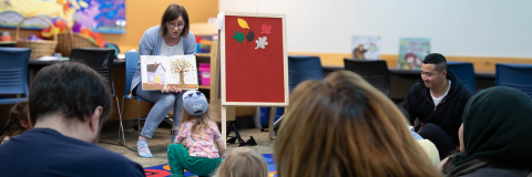 Young child sits on floor in front of Agata, a librarian, who is sitting on chair reading a picture book during storytime. 